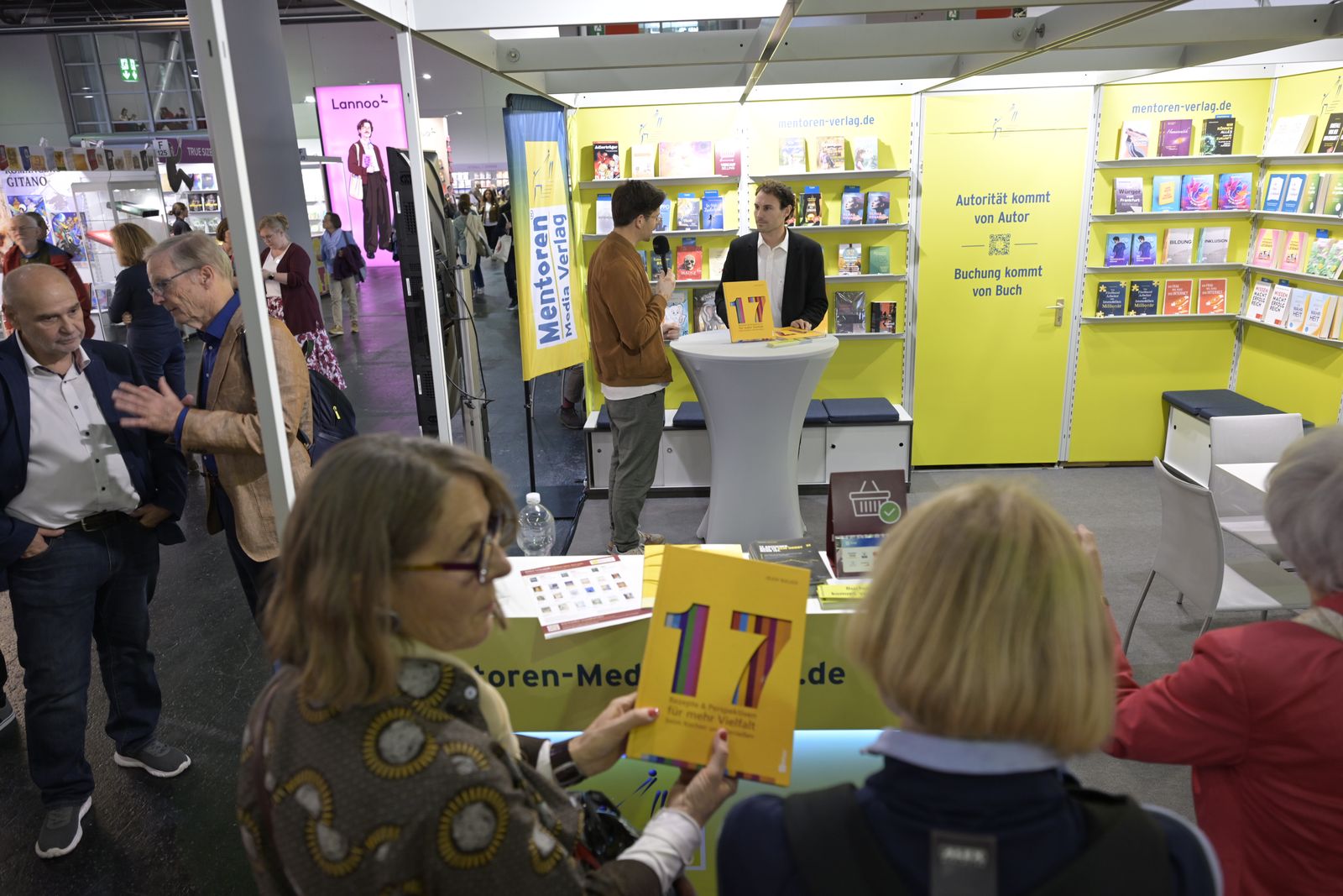 Menschen kommen am Messestand ins Gespräch, im Vordergrund das Buch „17“